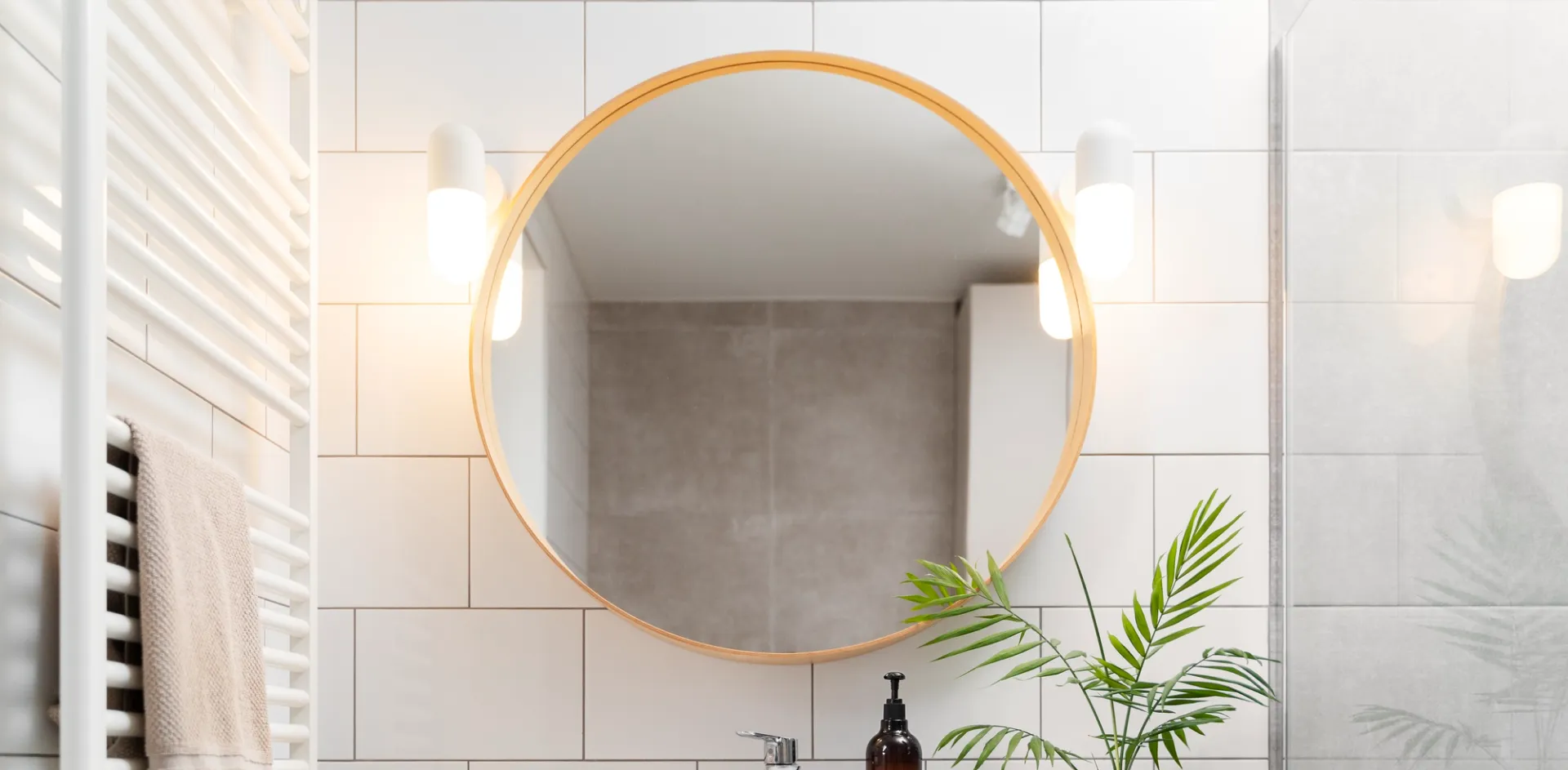 Round timber-framed bathroom mirror between two wall lights on white tiles, with a glass shower screen to the right