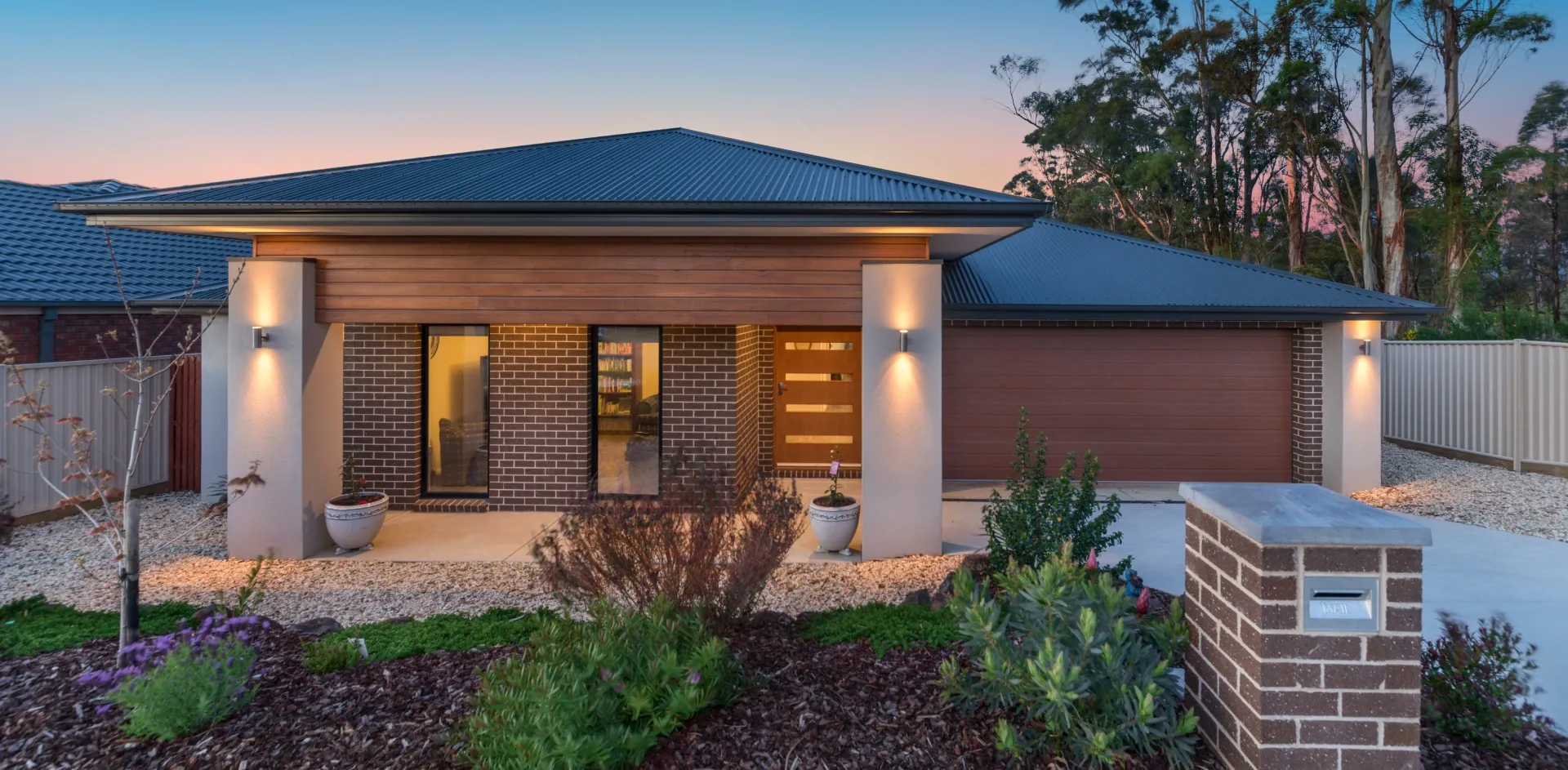 Modern brick home at dusk with tall black aluminium windows lit from inside beside a timber front door
