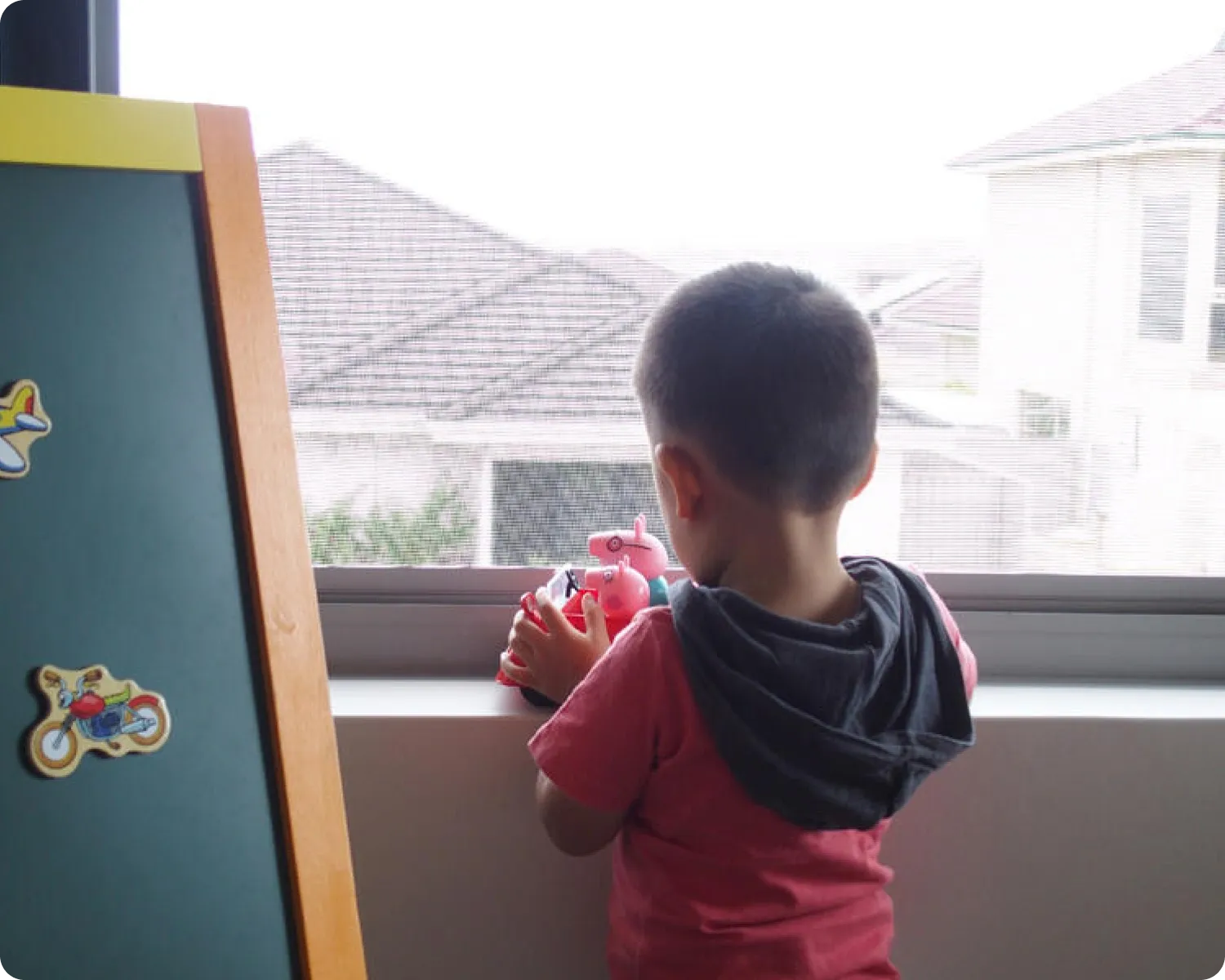 Young child holding a toy and leaning on the sill of a mesh-screened window, looking out at neighbouring rooftops