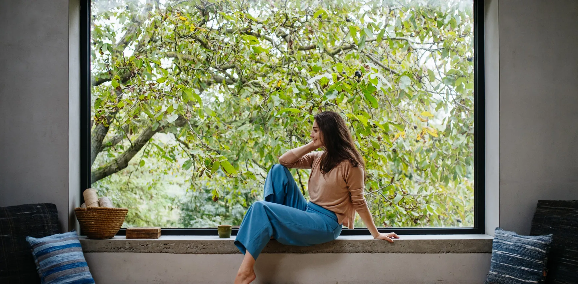Woman sitting on a concrete window ledge in front of a large fixed picture window with a slim black frame, looking out on a leafy tree