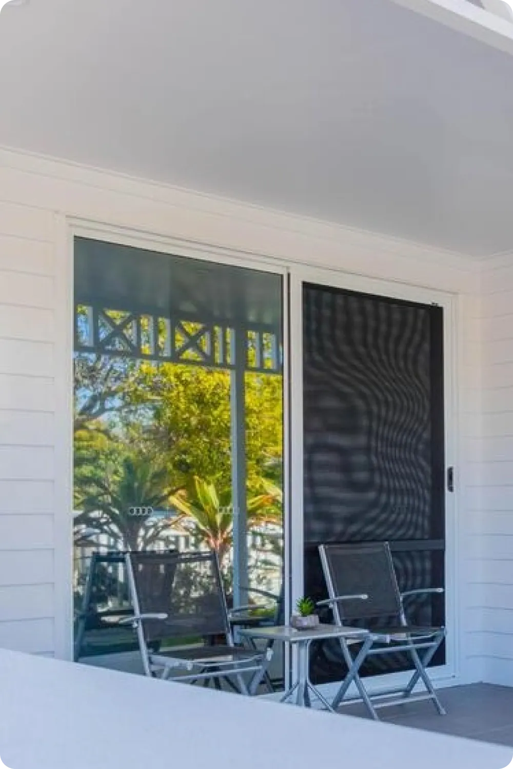 White aluminium sliding glass door with a black mesh safety screen door on a weatherboard verandah, with two outdoor chairs in front