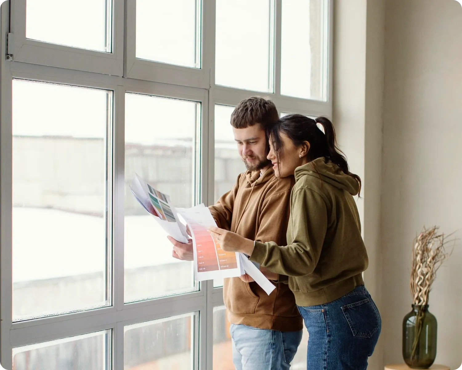 A couple comparing printed colour sample sheets beside a tall white-framed multi-pane window